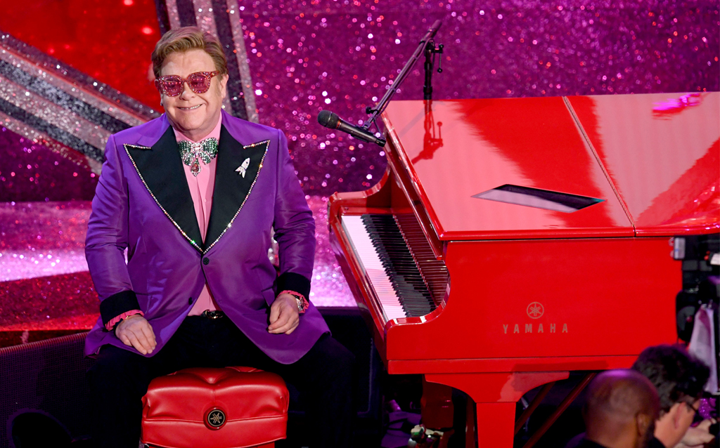 Sir Elton John wore a custom Gucci purple two button peak lapel tuxedo jacket with black contrast lapel and crystal trim with a pink shirt with ruffle detail, black tuxedo trousers, pink and green crystal bow tie, multicolor slip on sneakers and pink sunglasses with crystals.  